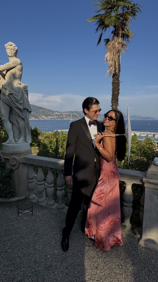 A couple in black-tie wedding-guest attire on a coastal terrace